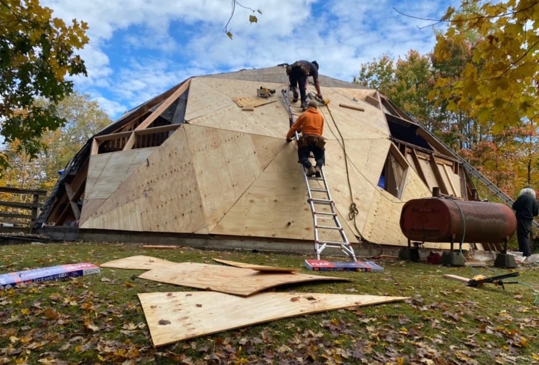 Geodesic dome under construction with plywood sheathing and roofing crew installing panels